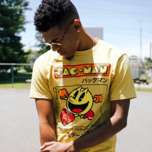 Casually dressed young man wearing a Pac-Man t-shirt, looking down outdoors under bright sunlight.